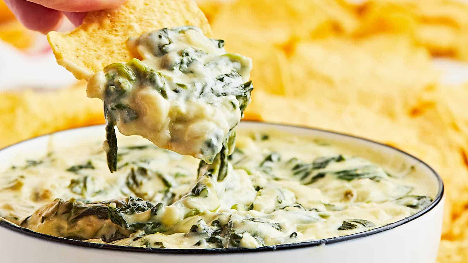 A person dipping a tortilla chip into a bowl of spinach dip.
